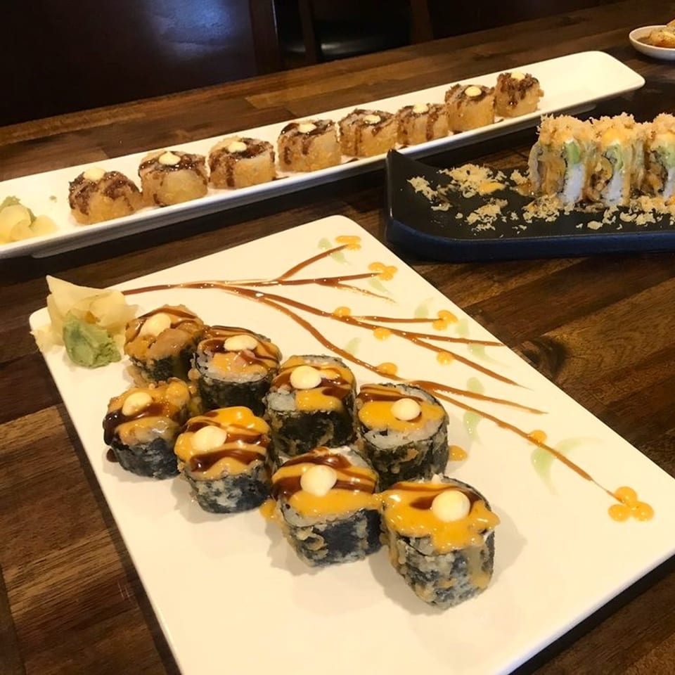 Three plates of sushi rolls, including deep-fried and sauced varieties, on a wooden table.